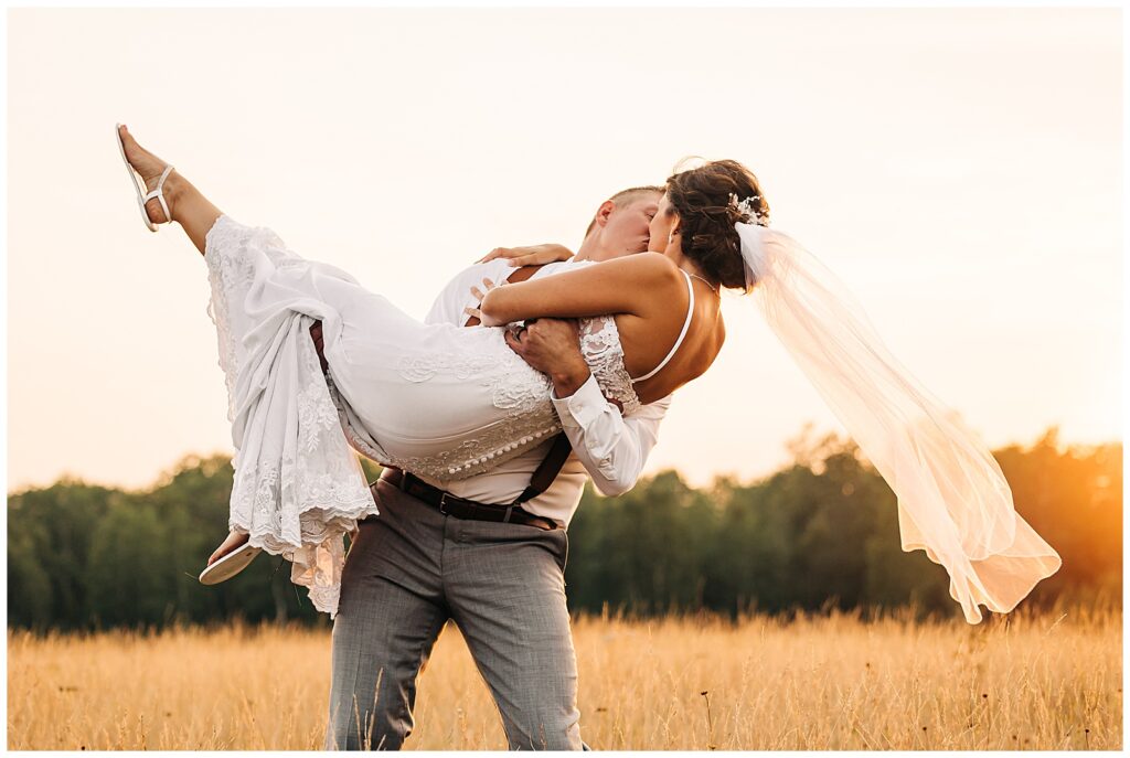 groom picking up and holding bride and kissing her in a field at sunset