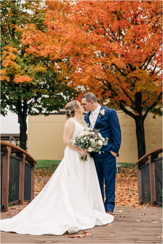 bride and groom kissing on bridge in front of fall trees at the Minnesota Discovery Center